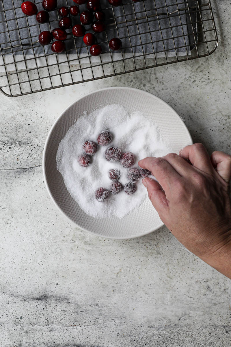 1 hand rolling the cranberries in sugar inside a small bowl with some cranberries on top of a wire rack on the top of the frame.