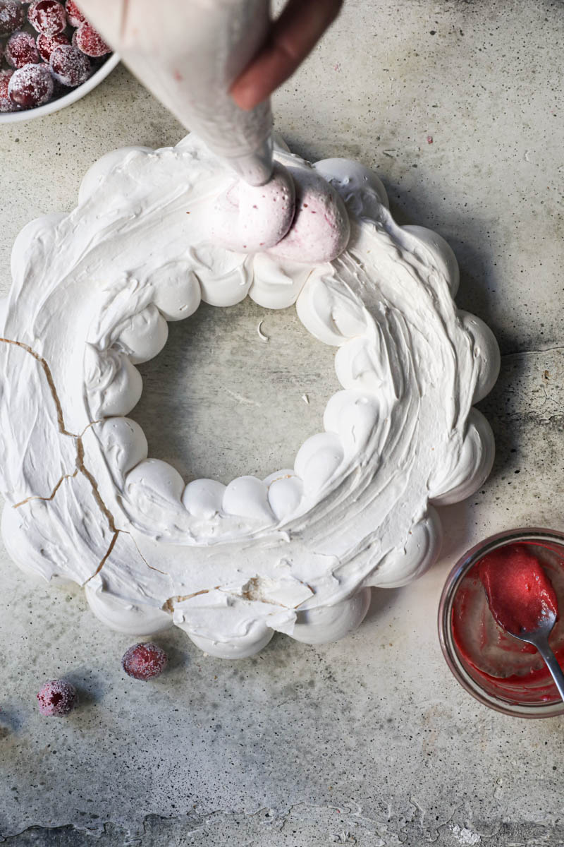 2 hands piping out the cranberry curd cream onto the Swiss meringue pavlova wreath.