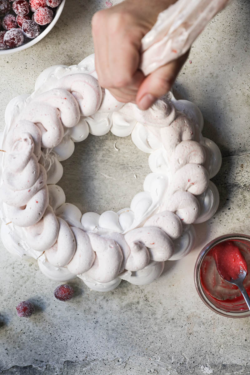 2 hands piping out the cranberry curd cream onto the Swiss meringue pavlova wreath.