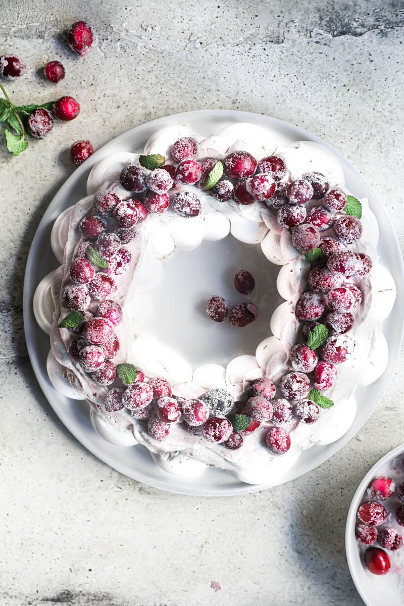 The Christmas Pavlova Wreath topped with cranberry curd Chantilly and sugar cranberries on a white plate with some cranberries on the side.