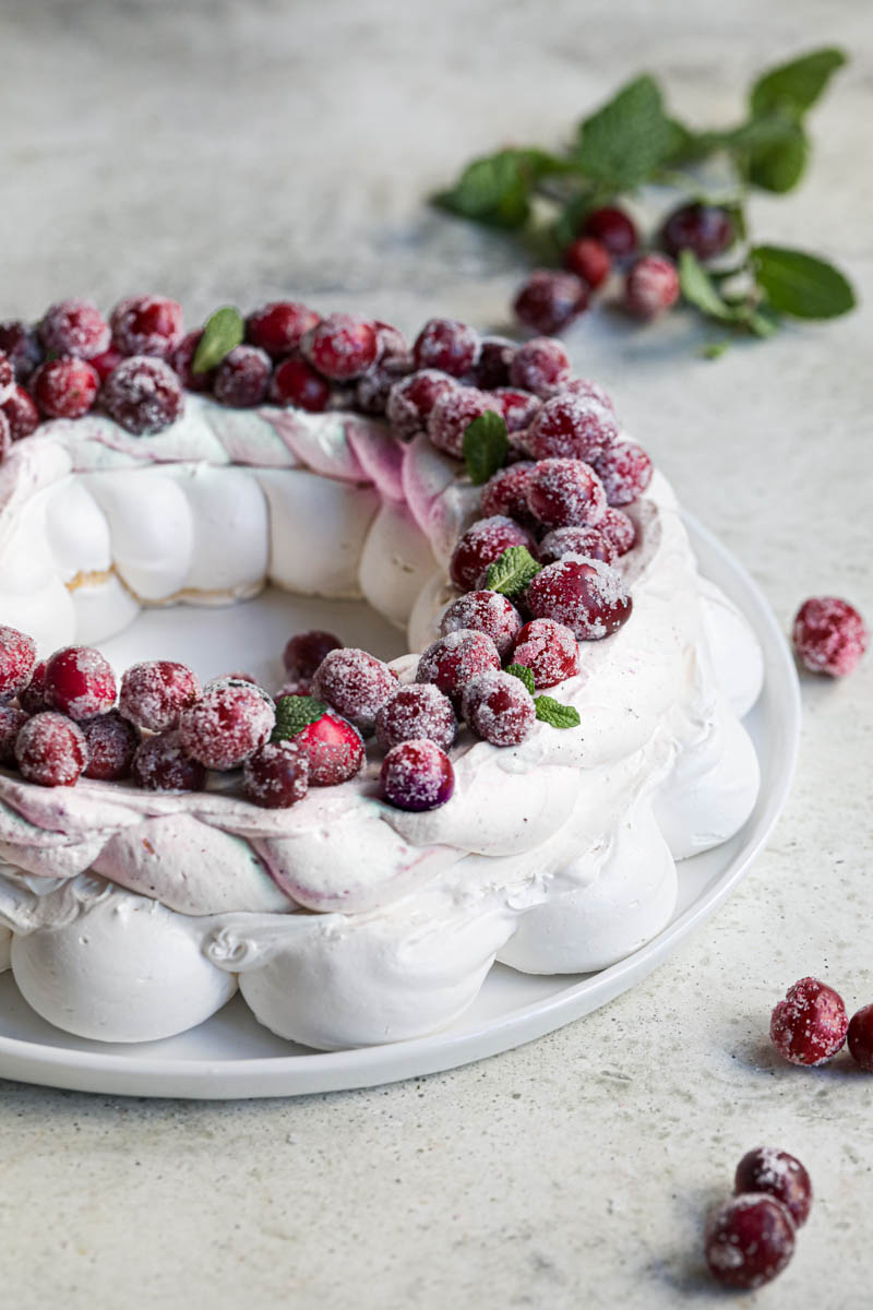 Closeup of the Christmas Pavlova Wreath topped with cranberry curd Chantilly and sugar cranberries on a white plate with some cranberries on the side, as seen from the side.