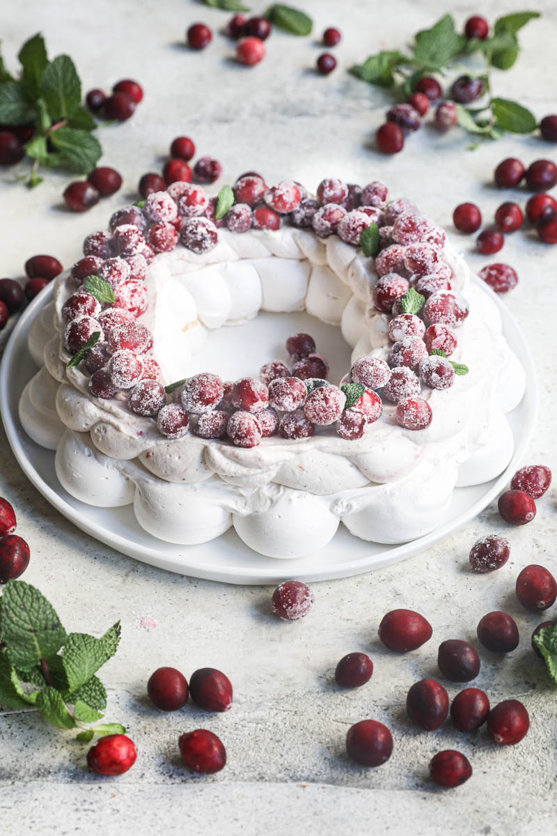 The Christmas pavlova wreath topped with sugar cranberries on a white plate with cranberries and mint leaves all around.