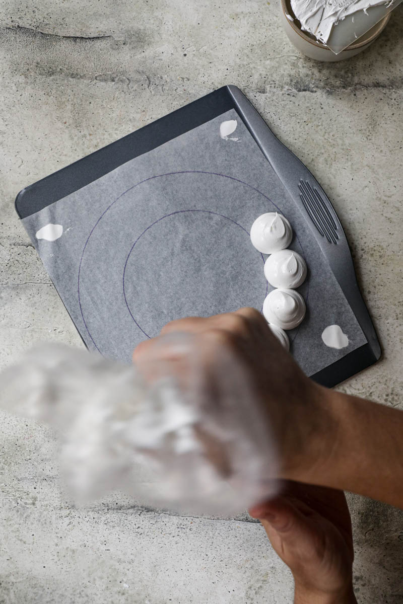 2 hands piping out the meringue onto a baking tray lined with parchment paper in the shape of wreath.