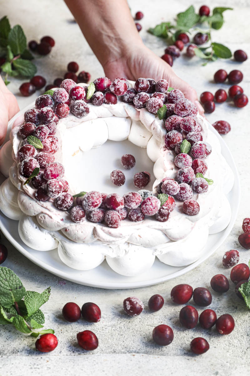 2 hands holding the Christmas pavlova wreath topped with sugar cranberries with cranberries and mint leaves all around.