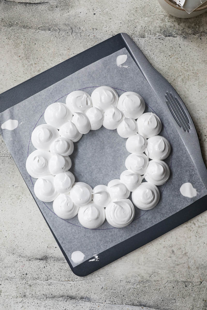 2 hands piping out the meringue onto a baking tray lined with parchment paper in the shape of wreath.