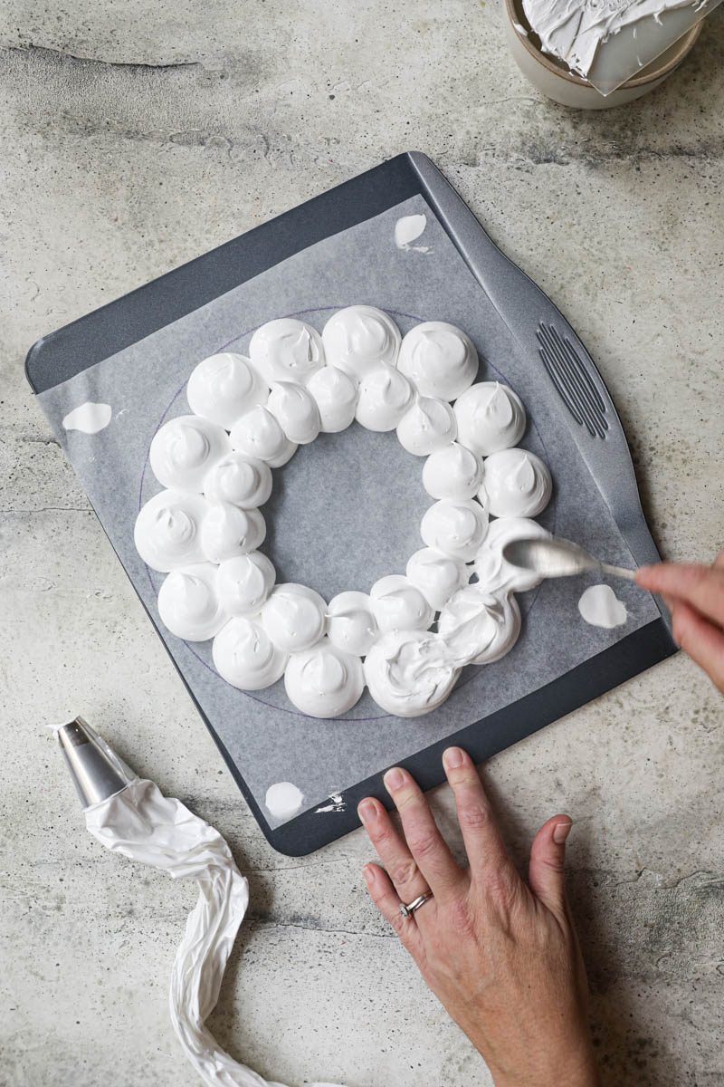 1 hand smoothing out the pavlova wreath on top of a baking tray lined with parchment paper using a spoon.