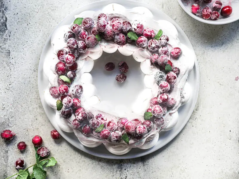 The Christmas Pavlova Wreath topped with cranberry curd Chantilly and sugar cranberries on a white plate with some cranberries on the side.