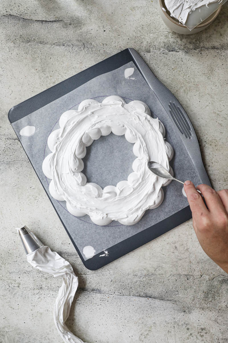 1 hand smoothing out the pavlova wreath on top of a baking tray lined with parchment paper using a spoon. 