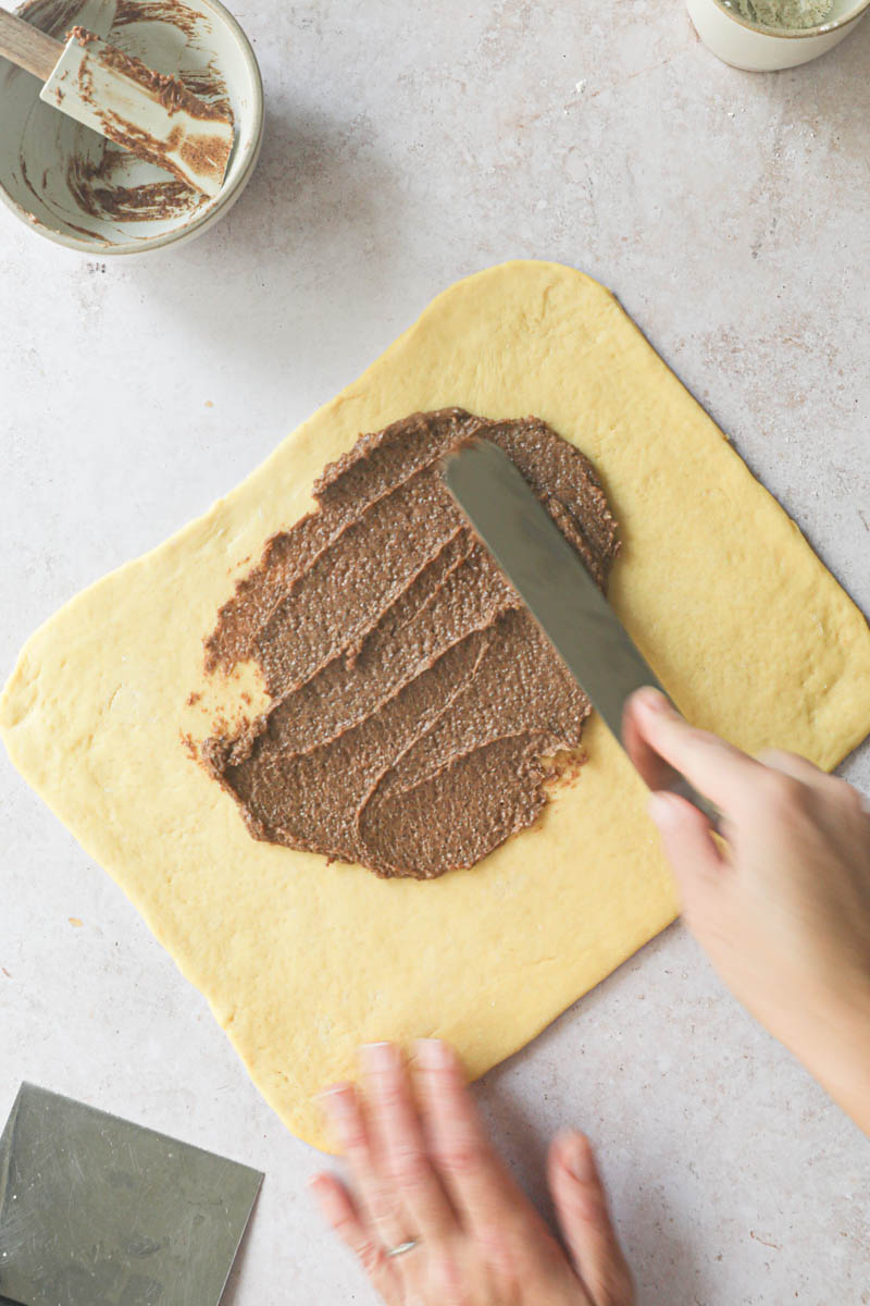 One hand spreading the cinnamon roll filling onto the rectangular brioche dough.