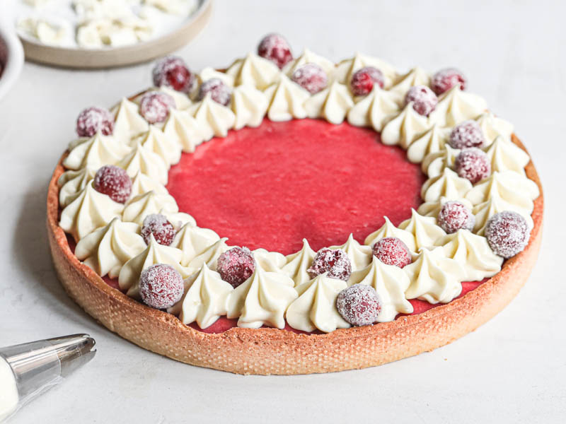 The cranberry curd tart topped with chocolate ganache kisses seen from the side with a small plate in the left-hand side and a piping tip on the bottom left corner.