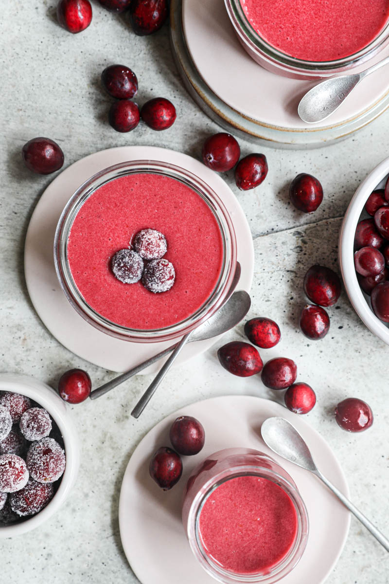 The cranberry curd inside a glass jar placed on top of plate with 2 spoons on the side and some sugared cranberries on top, seen from above.