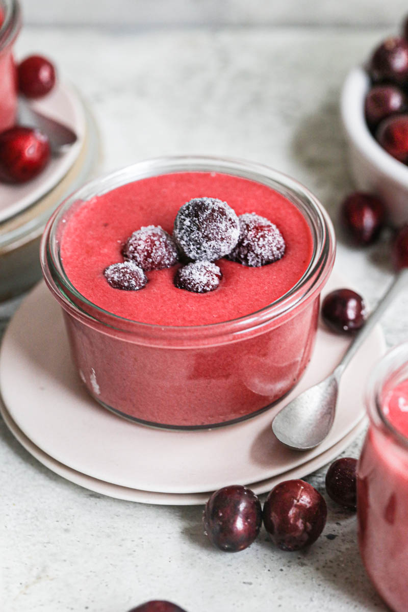 The cranberry curd inside a glass jar placed on top of plate with 2 spoons on the side and some sugared cranberries on top, some fresh cranberries around and a plate blurry in the back.