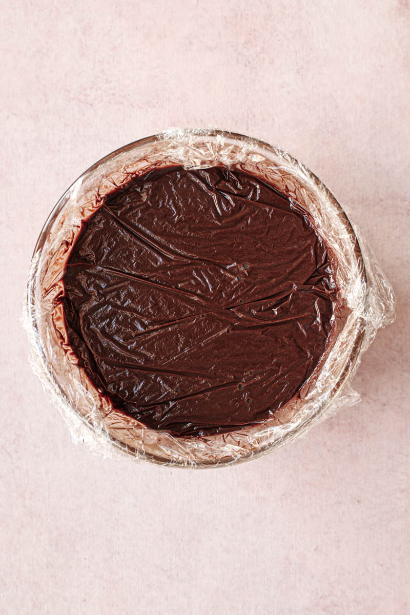 The creamy dark chocolate ganache inside a glass bowl covered in saran wrap.