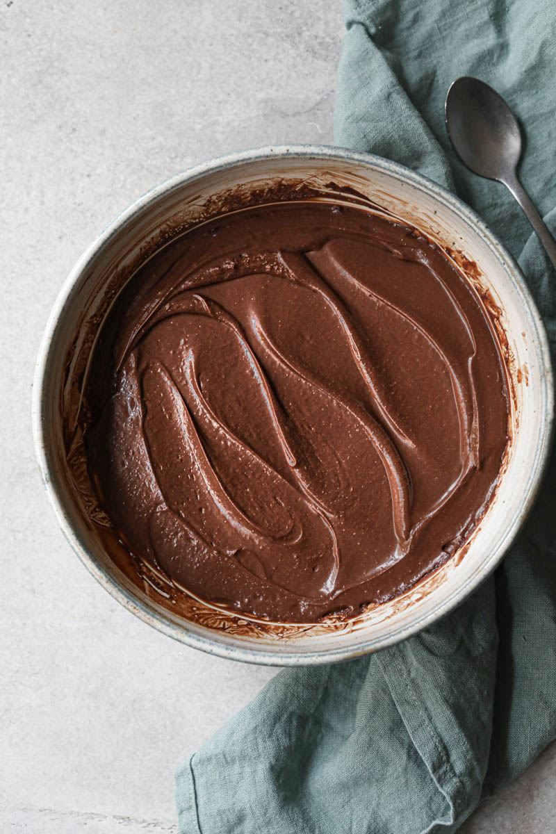 The creamy dark chocolate ganache after it has set inside a grey bowl with a blue linen and a spoon on the side.