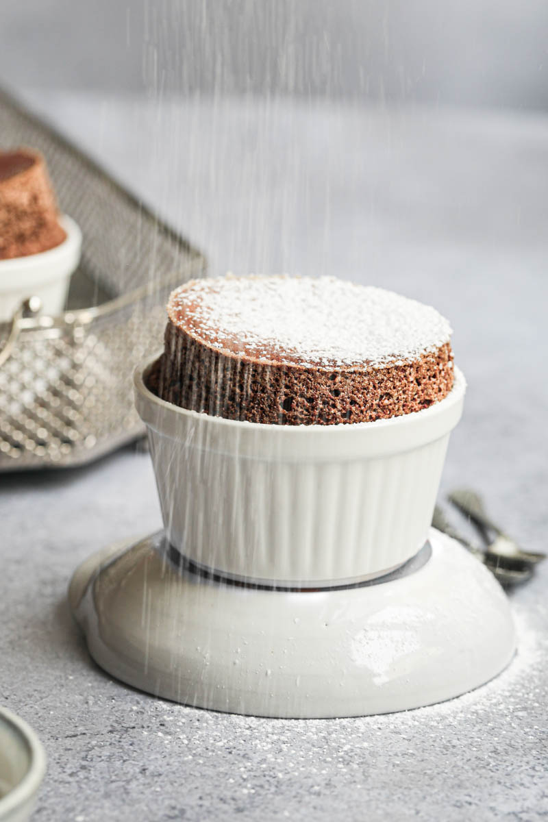 The chocolate souffle on top of a grey bowl being sprinkled with powdered sugar, with 2 spoons on the side, and a tray with the other dark chocolate souffle blurry in the back.