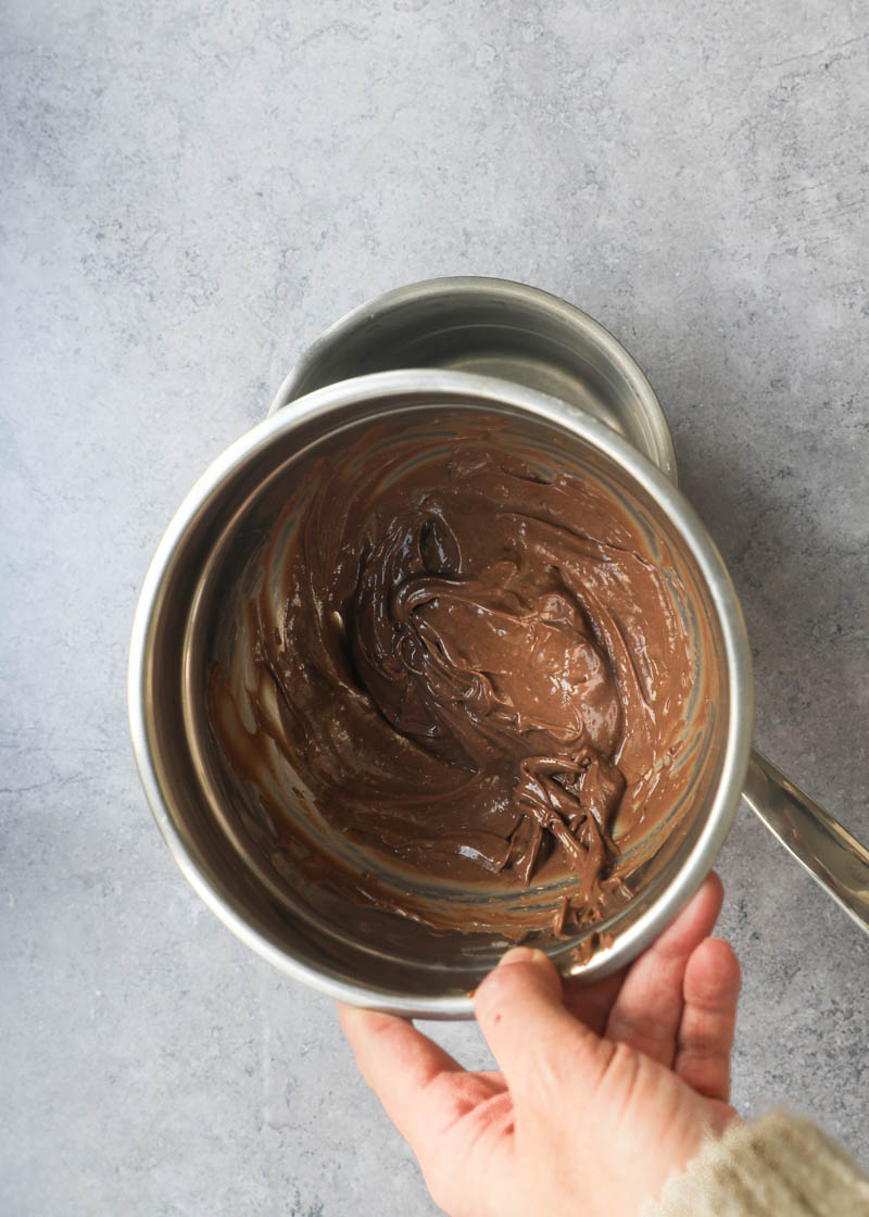 One hand holding a bowl with the chocolate pastry cream and a pan full of water underneath.