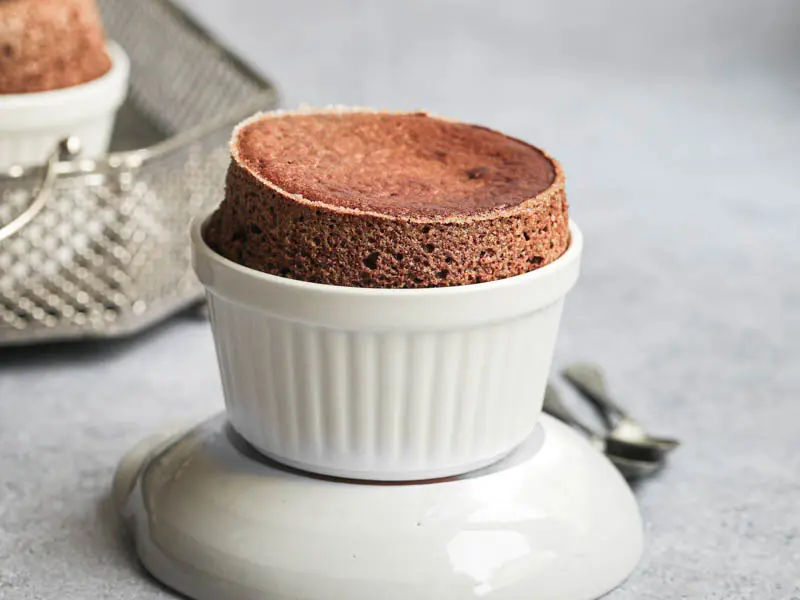 The chocolate souffle on top of a grey bowl with 2 spoons on the side, and a tray with the other dark chocolate souffle blurry in the back.