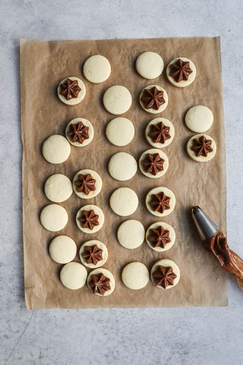 The alfajores cookies on top a piece of parchment paper, turned over and filled with dulce de leche with a piping bag on the side.