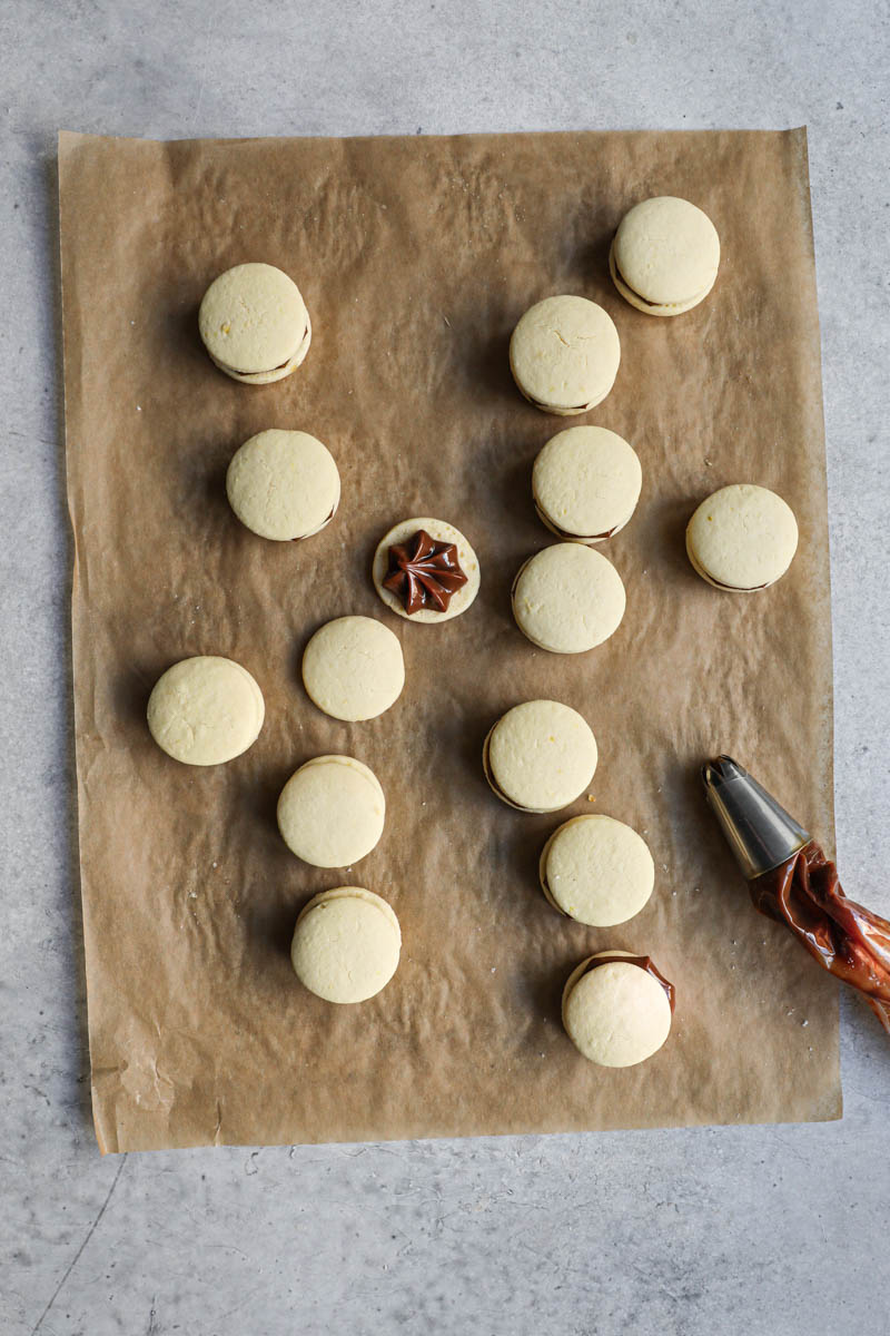 The alfajores cookies on top a piece of parchment paper, turned over, filled with dulce de leche and covered with a second corn-starch cookie, with a piping bag on the side.