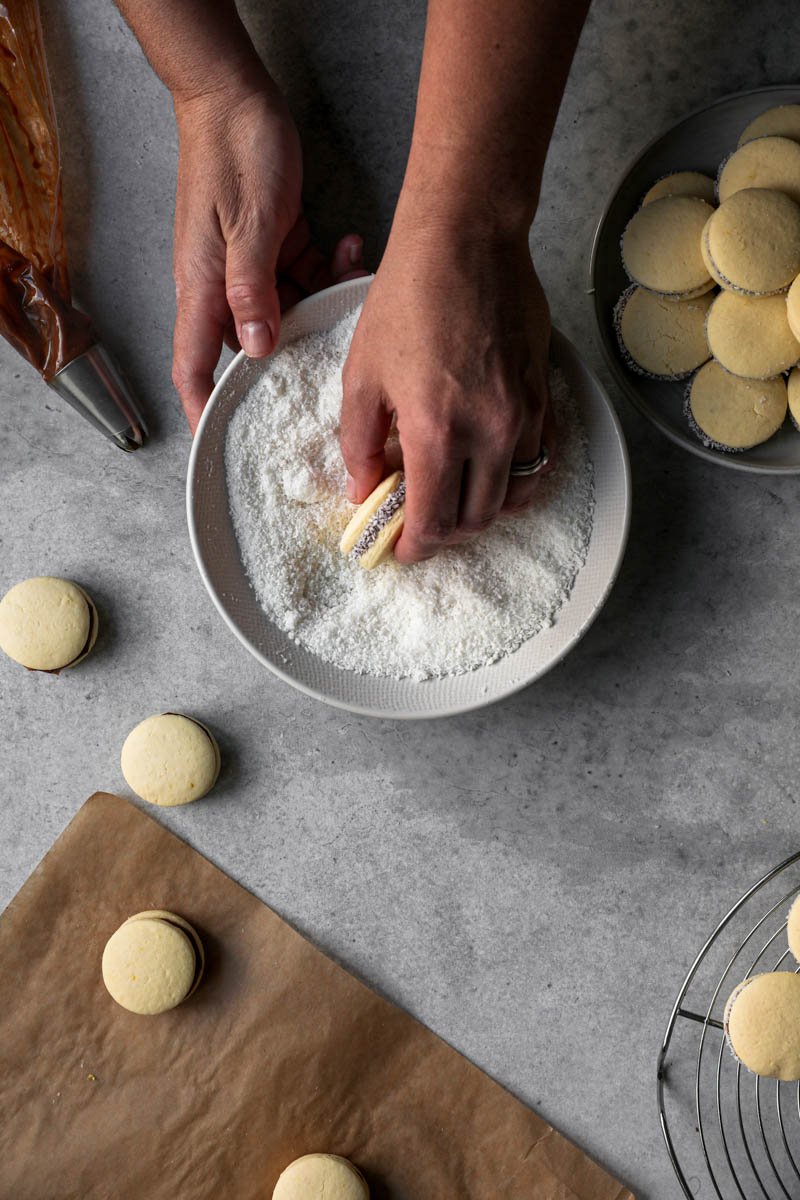 One hand rolling one alfajor inside a bowl filled with grated coconut, with some alfajores around the bowl.