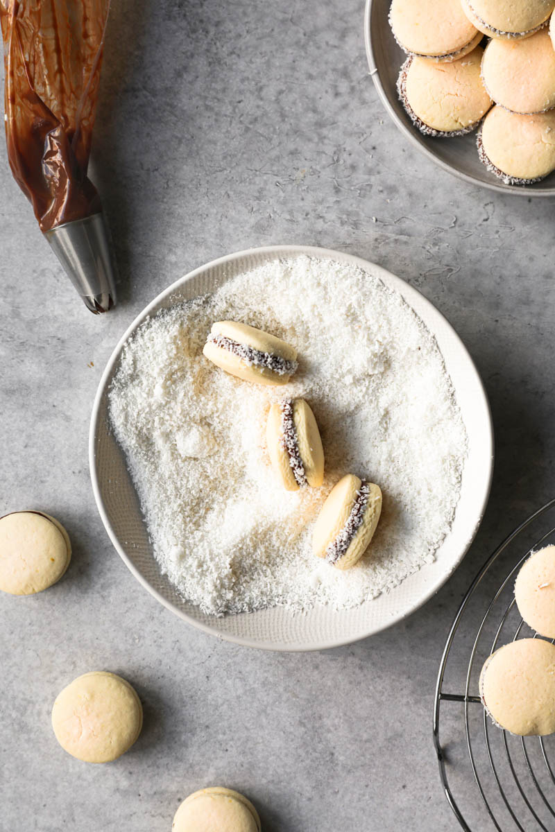 3 alfajores cookies inside a bowl with shredded coconut with some alfajores around the bowl.