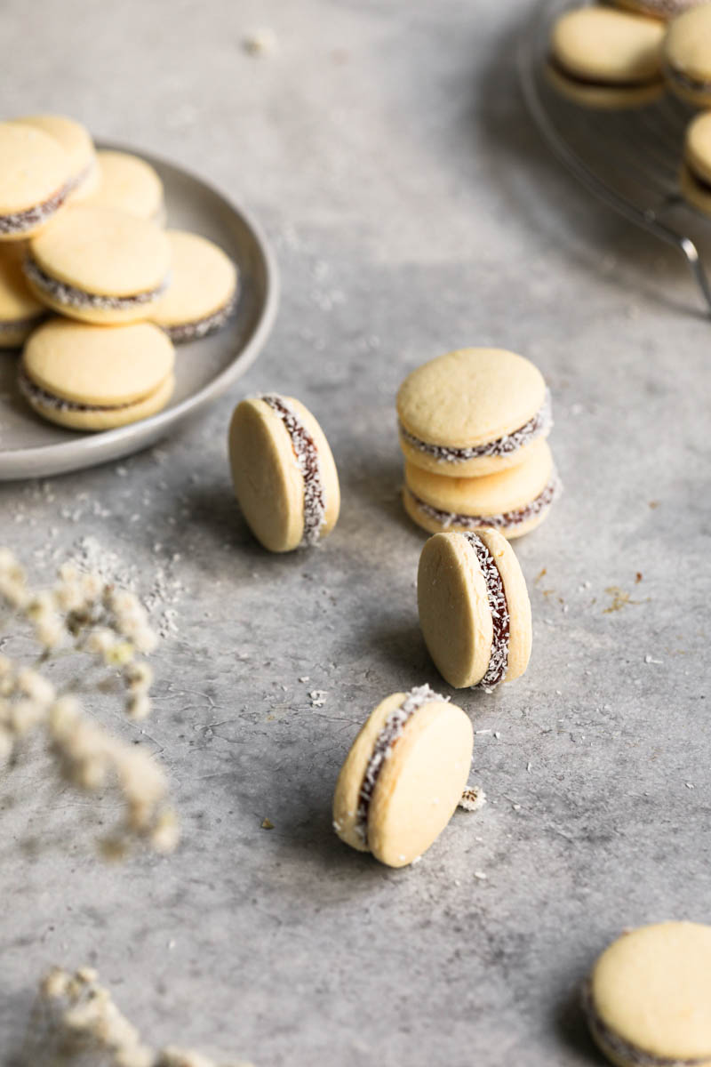 Some alfajores on top of a grey work surface with a plate full of dulce de leche alfajores on the left-hand side of the frame.