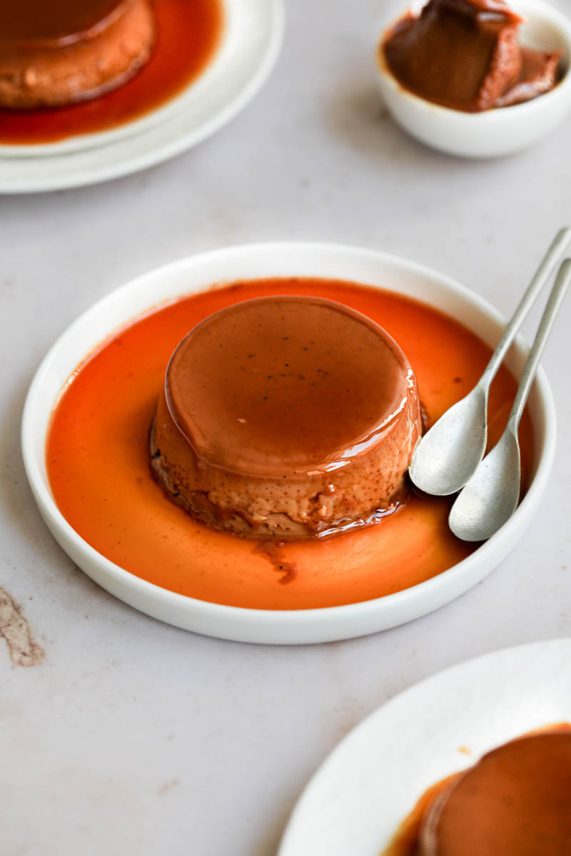 Serving the mini-individual dulce de leche flan with milk: one dulce de leche flan on a small white plate with 2 spoons on the side. 