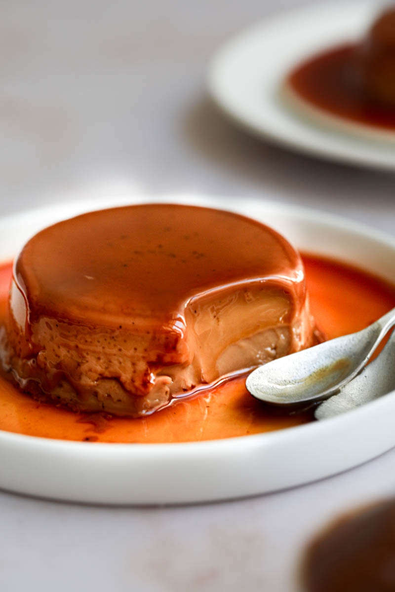 Closeup shot of one mini-individual dulce de leche flan on a white plate with a spoon on the side with one bite missing and a plate in the background.