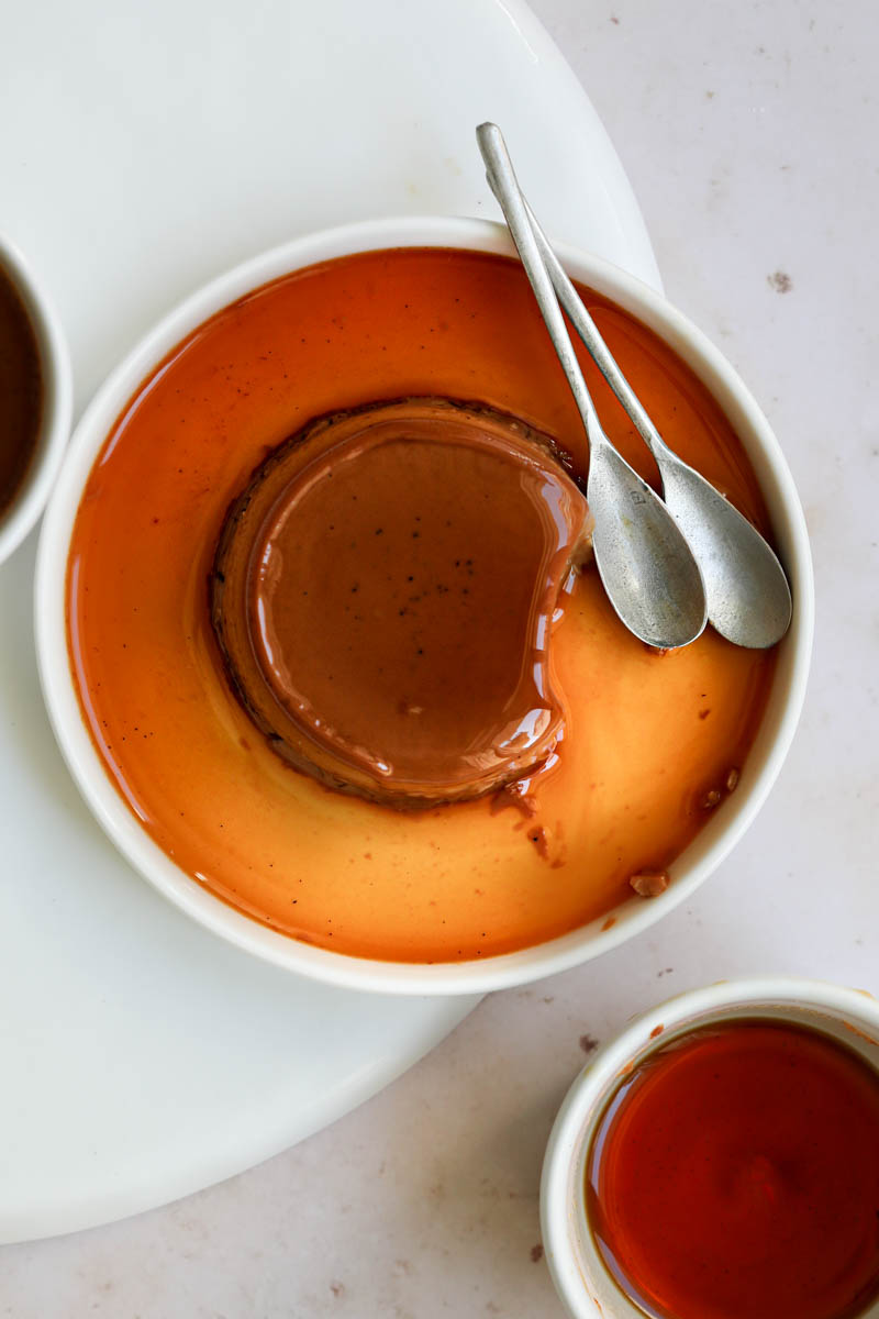 One mini-individual dulce de leche flan on a white plate with a spoon on the side with one bite missing and an empty ramekin on the bottom right corner.