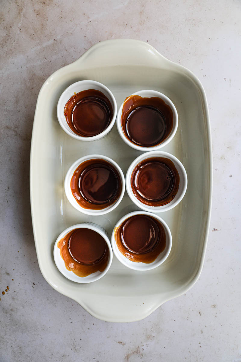 The ramekins lined with caramel for the dulce de leche flan inside a baking tray.