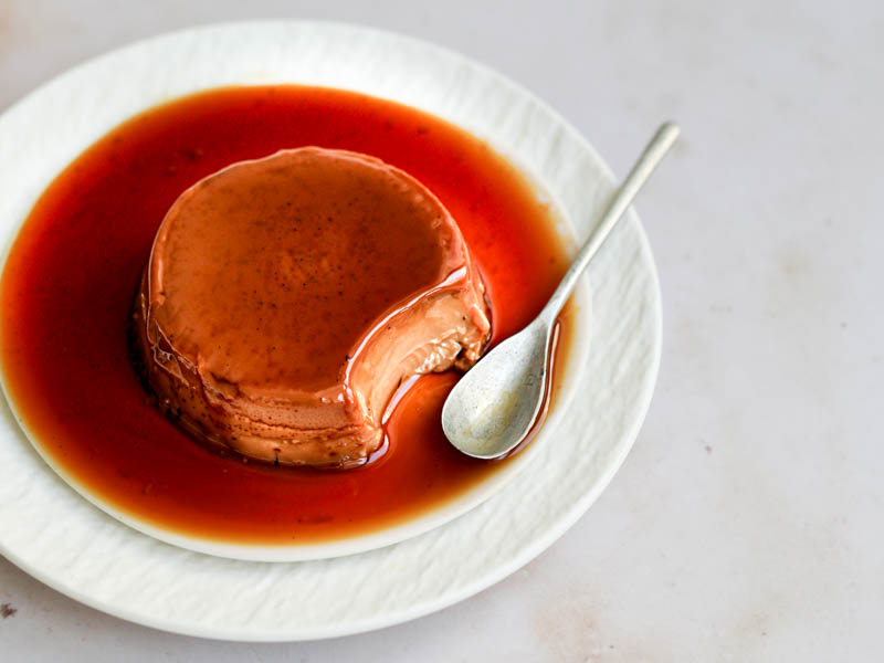 One mini-individual dulce de leche flan on a pink plate placed over a larger white plate with a spoon on the side. One bite is missing.