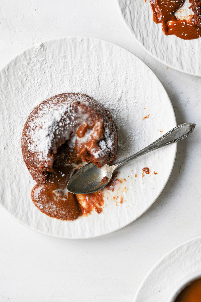 The dulce de leche lava cake with a scoop of melted ice cream and a spoon on the side seen from above with the edges of two plates seen on each corner.