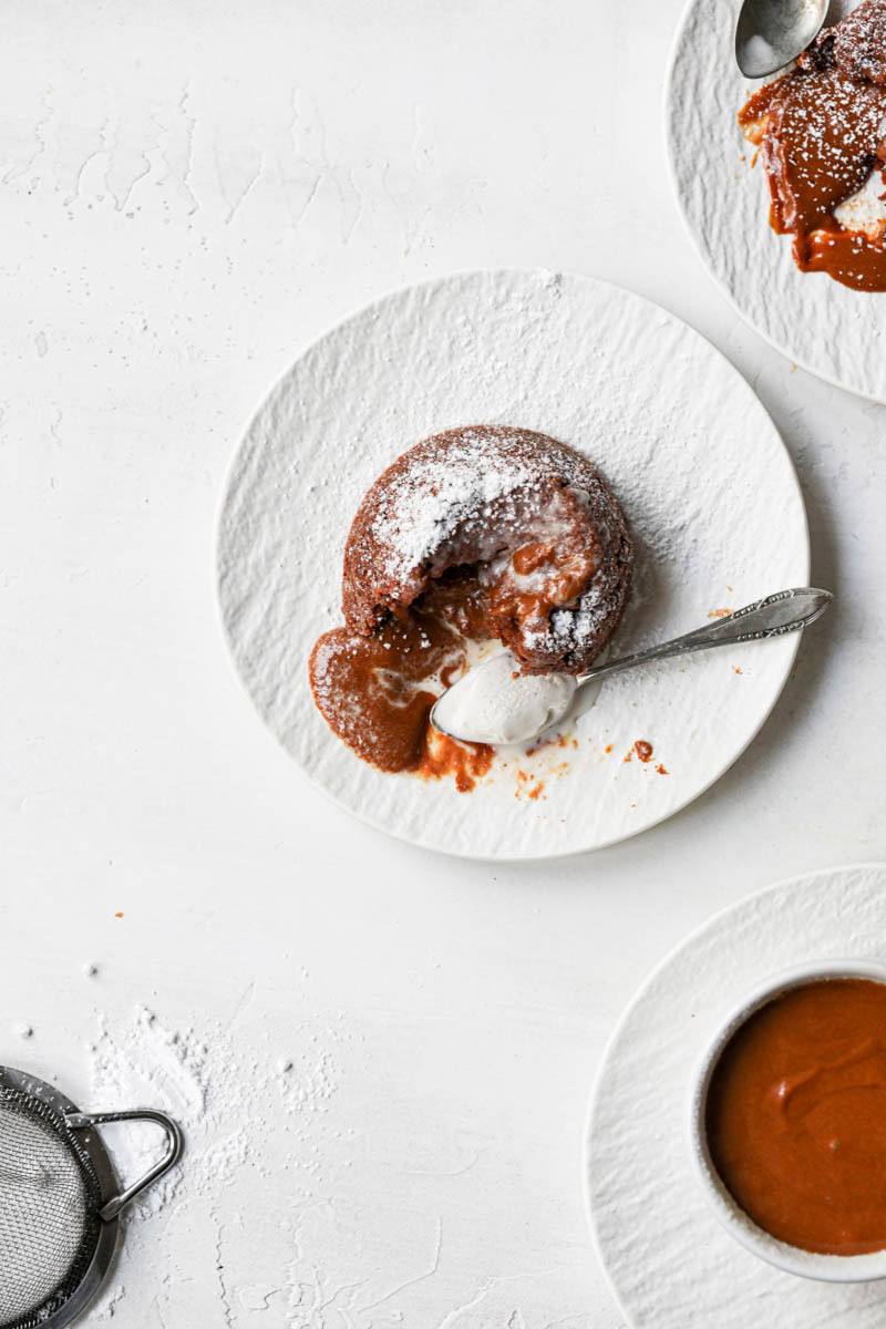 The dulce de leche lava cake with a scoop of melted ice cream and a spoon on the side seen from above with a small strainer on the bottom left corner and a plate with a smashed cake on the top right corner.