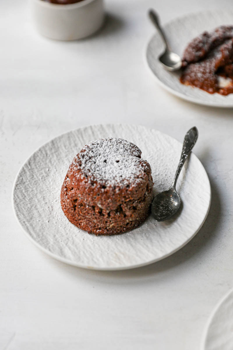 The whole dulce de leche lava cake on a white plate with spoon on the side and a plate in the back.