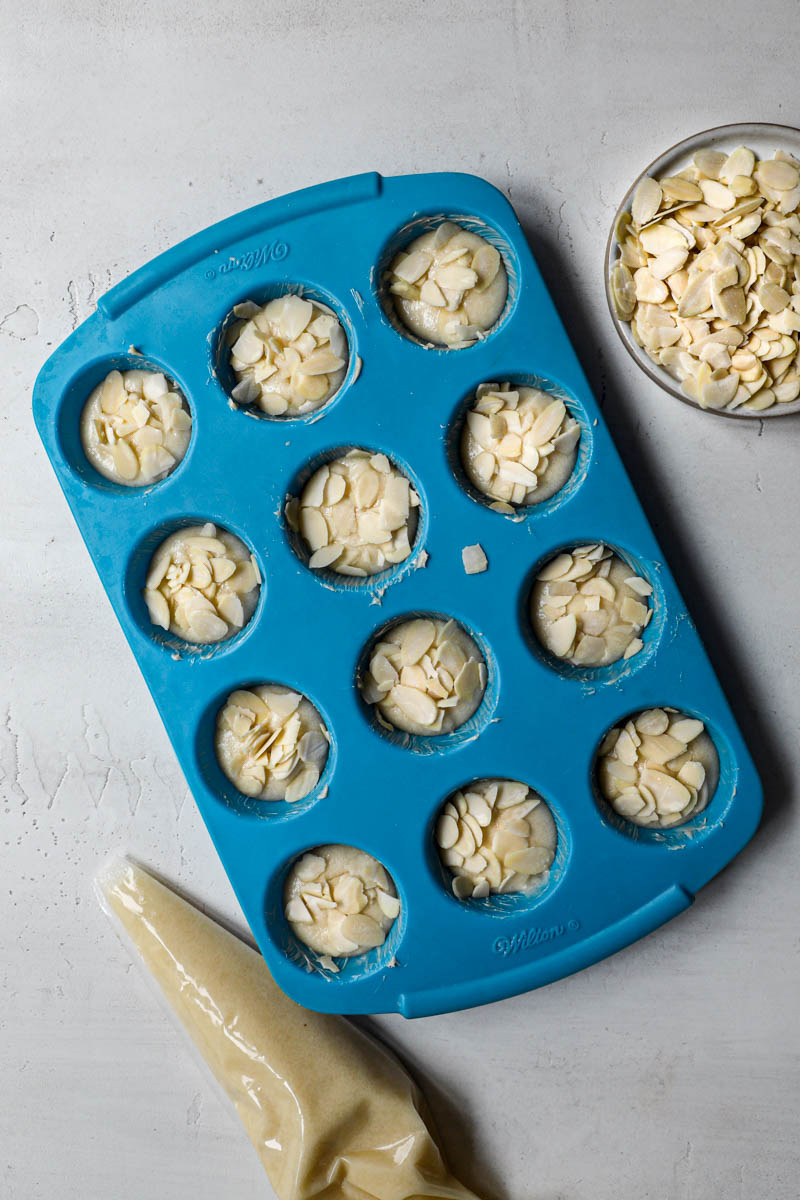 A blue mini muffin pan filled with the French almond cakes with a piping bag on the side and a plate holding sliced almonds on the side.