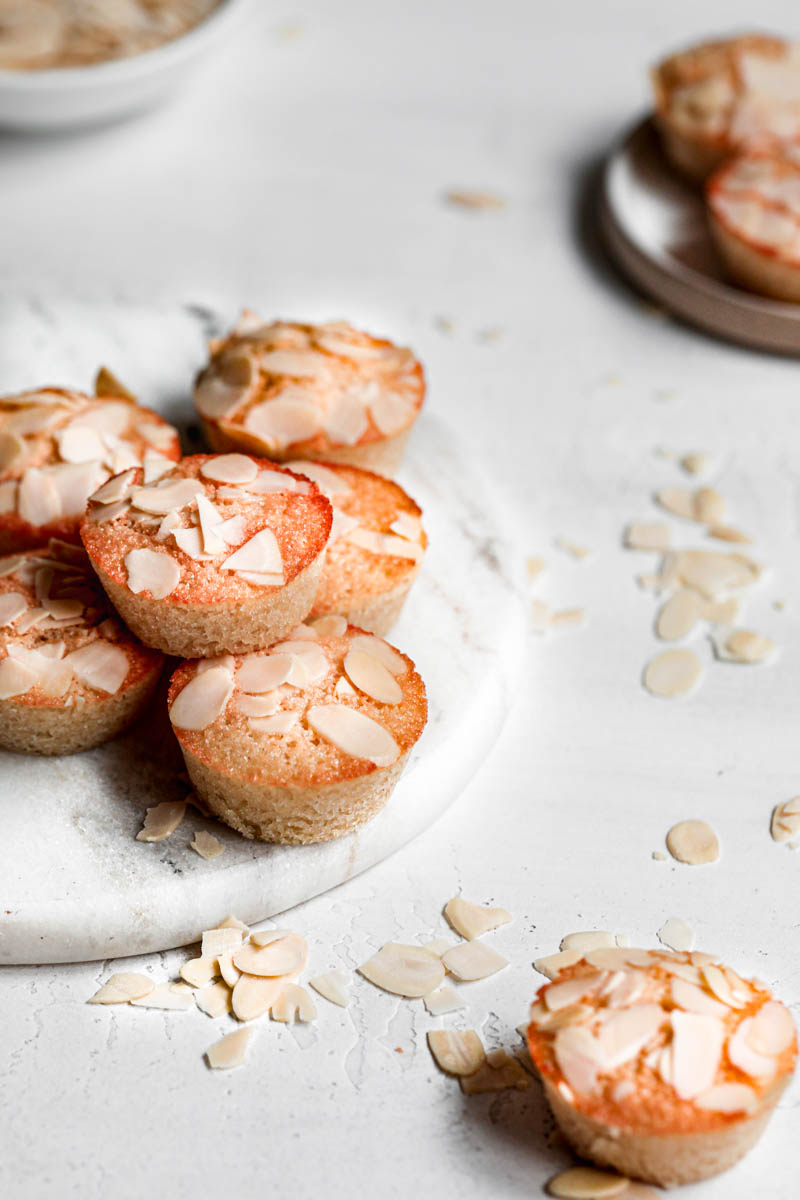 Stacked almond financier cakes on top of marble platter with some small financiers around it seen from the side.