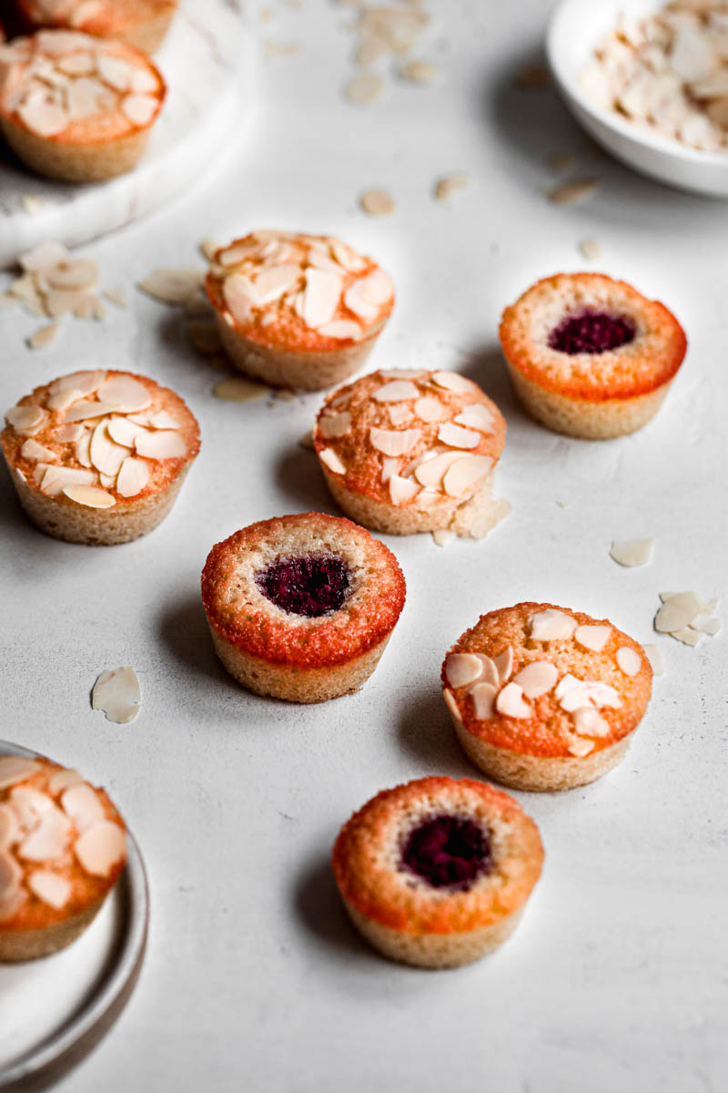 Almond financier cakes and raspberry financier cakes randomly placed on top of a white surface.