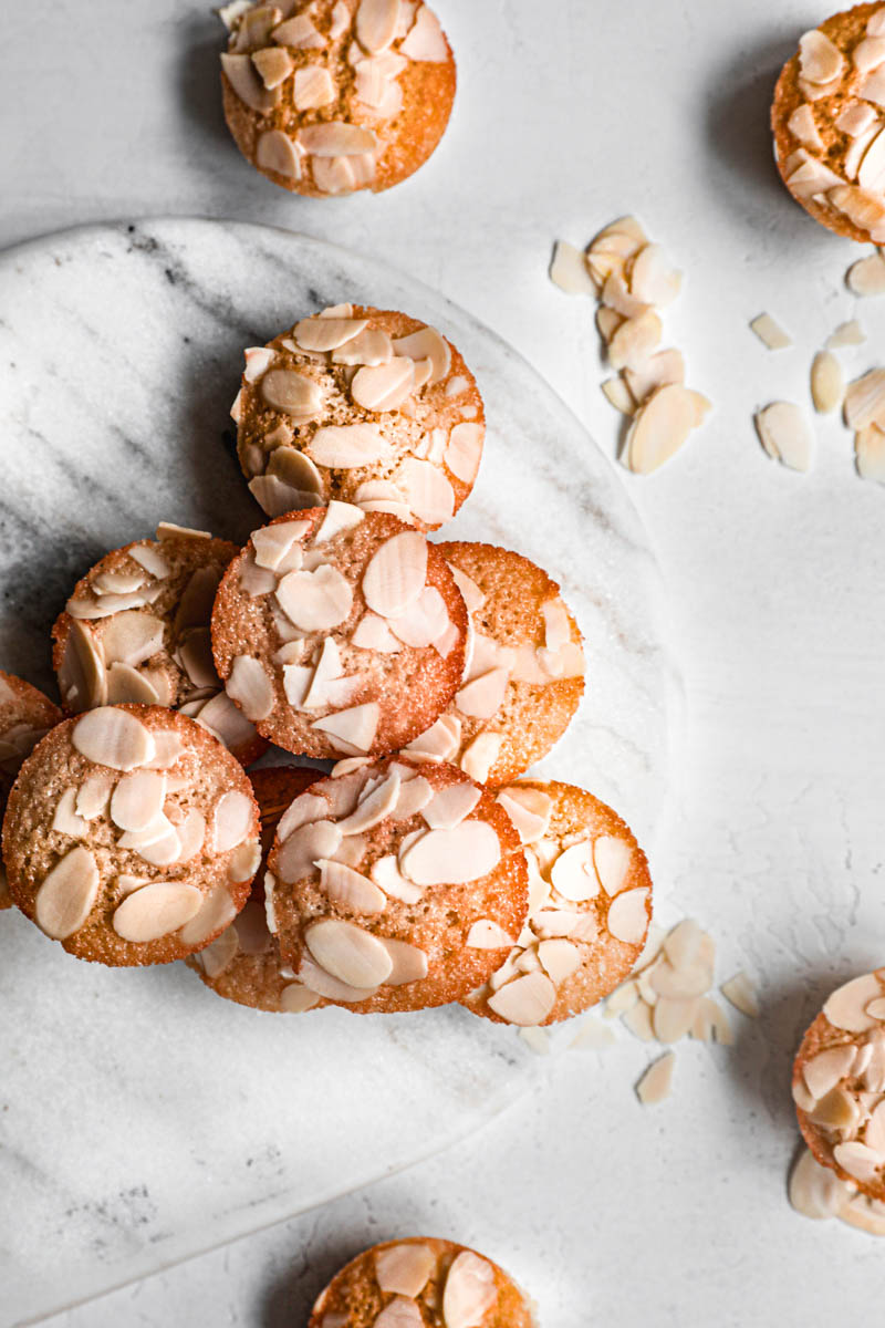 Stacked almond financier cakes on top of marble platter with some small financiers around it, as seen from above.