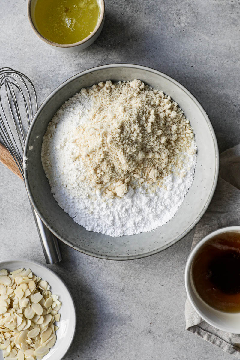 All the dry ingredients inside a grey bowl with some sliced almonds on a plate on top of the frame and egg whites in a bowl next to it.