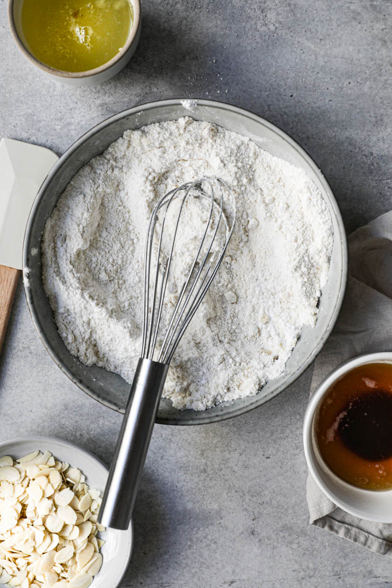 All the dry ingredients whisked together inside a grey bowl with some sliced almonds on a plate on top of the frame and egg whites in a bowl next to it and a whisk inside the bowl.