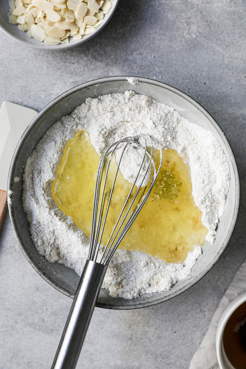 All the dry ingredients whisked together inside a grey bowl plus the egg whites, with some sliced almonds on a plate on top of the frame and egg whites in a bowl next to it and a whisk inside the bowl.