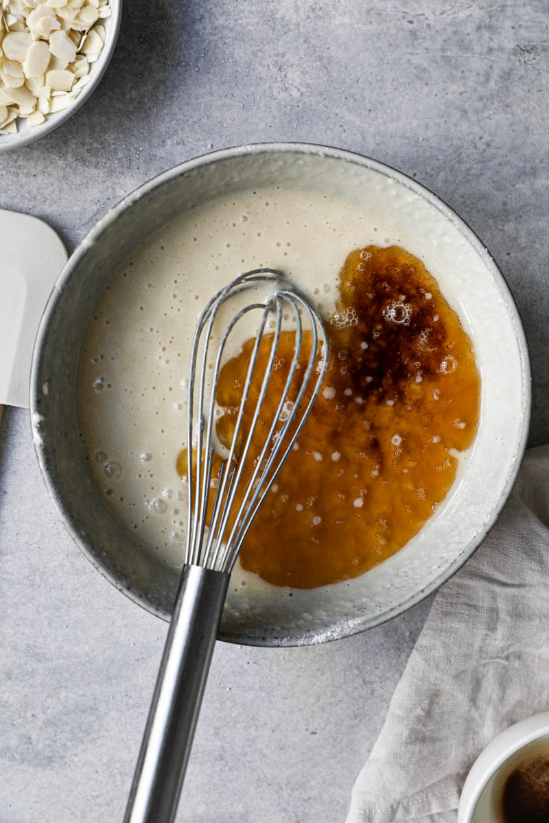 The financier cake batter in a grey bowl with a whisk inside it plus the added brown butter before mixing it in.