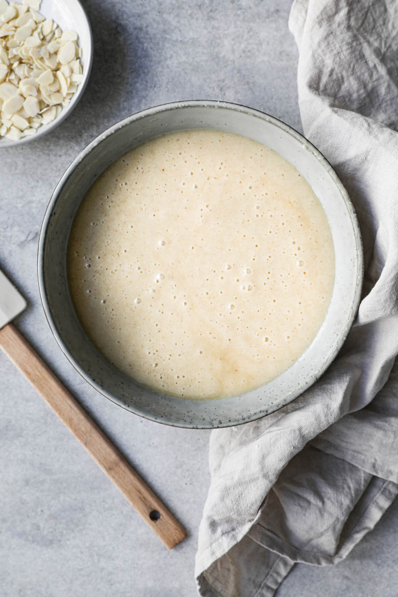 The financier cake batter in a grey bowl ready to be transferred to the pan with a rubber spatula on the side and beige linen underneath.