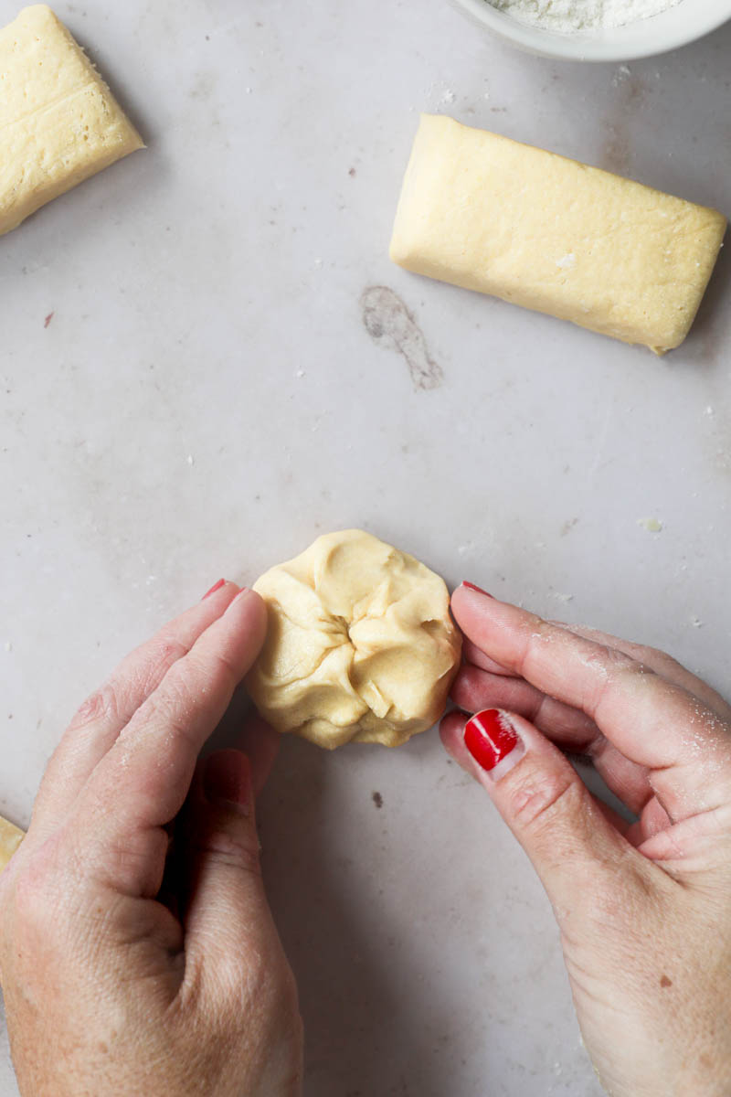 2 Hands shaping one piece of brioche bread dough into a tight ball with other pieces of dough around.