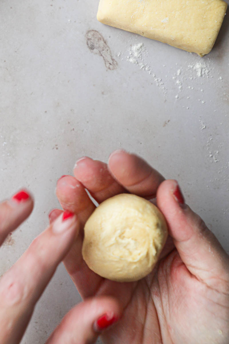2 Hands shaping one piece of brioche bread dough into a tight ball with other pieces of dough around.