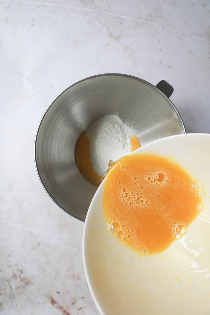 One hand pouring the mixture egg/mil/yeast on top of the flour in a bowl.