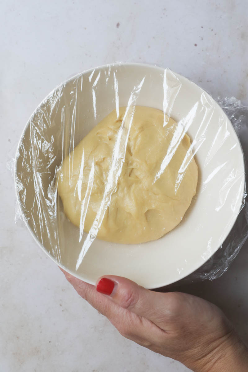 The French brioche bread dough ready inside a beige bowl ready for first rise, covered with saran wrap.