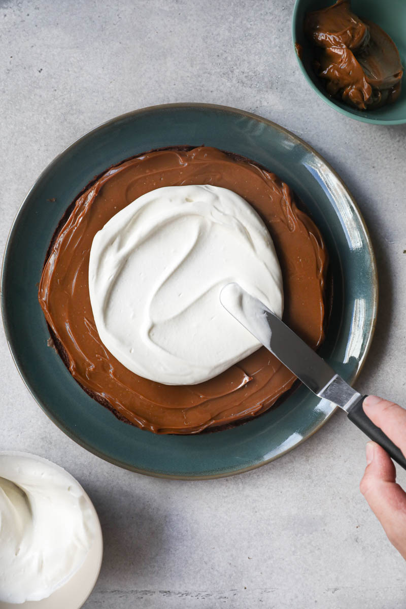 One hand spreading the whipped cream over the French chocolate cake with a small off set spatula.