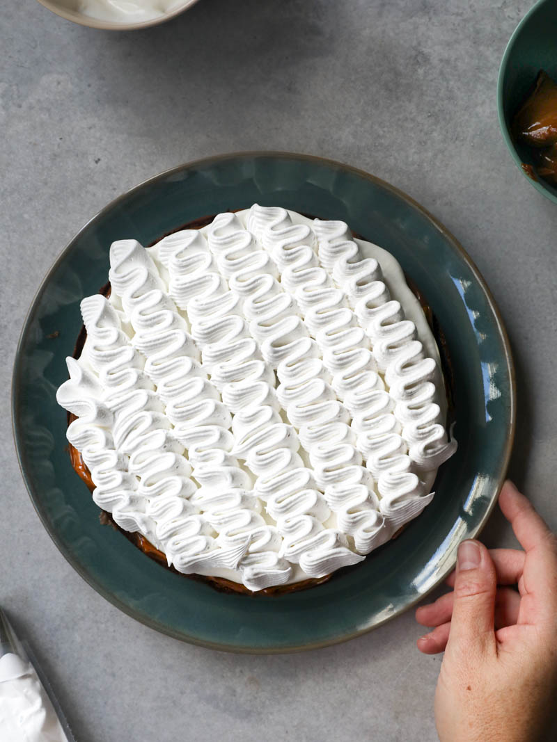 On hand the holding a blue plate with the French chocolate cake filled with whipped cream with a piping bag on the side and a bowl on top.