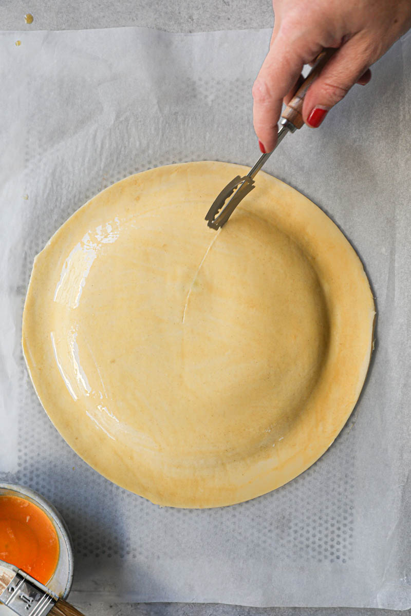 One hand scoring the French king cake with a scoring knife.